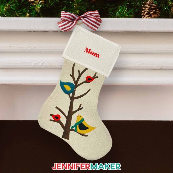 Create a DIY personalized Christmas stocking with sublimation with JenniferMaker's tutorial! A burlap stocking hangs from a white mantle in front of a fireplace. Pinecones and branches decorate the top of the mantle, and the stocking hangs from a striped bow. The stocking is off-white and has multicolored birds on a tree on the front, with the name "Mom" on the cuff.