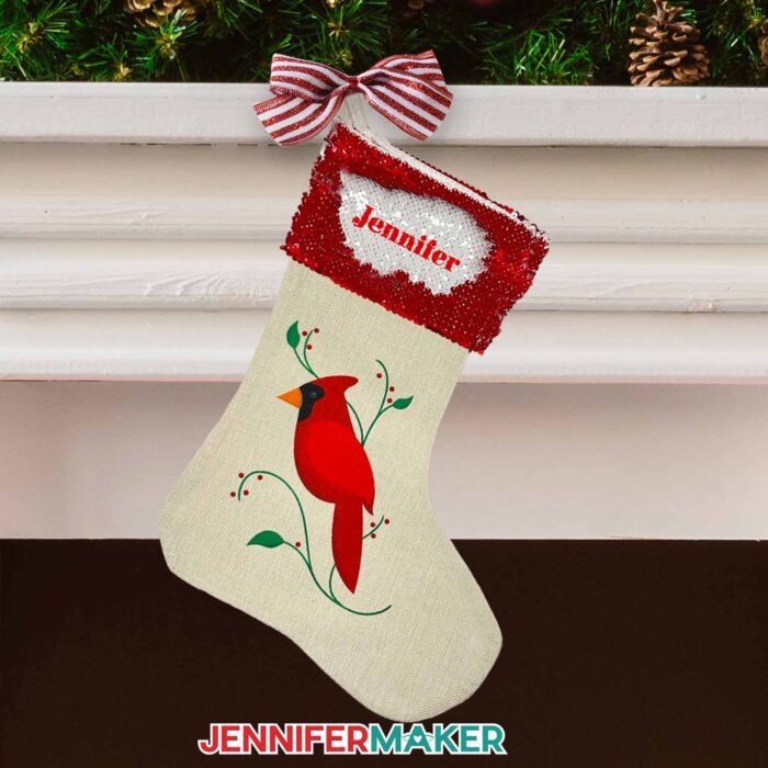 Create a DIY personalized Christmas stocking with sublimation with JenniferMaker's tutorial! A burlap stocking hangs from a white mantle in front of a fireplace. Pinecones and branches decorate the top of the mantle, and the stocking hangs from a striped bow. The stocking is off-white and has a cardinal bird on the front, with the name "Mom" on the red and white flippable sequin cuff.