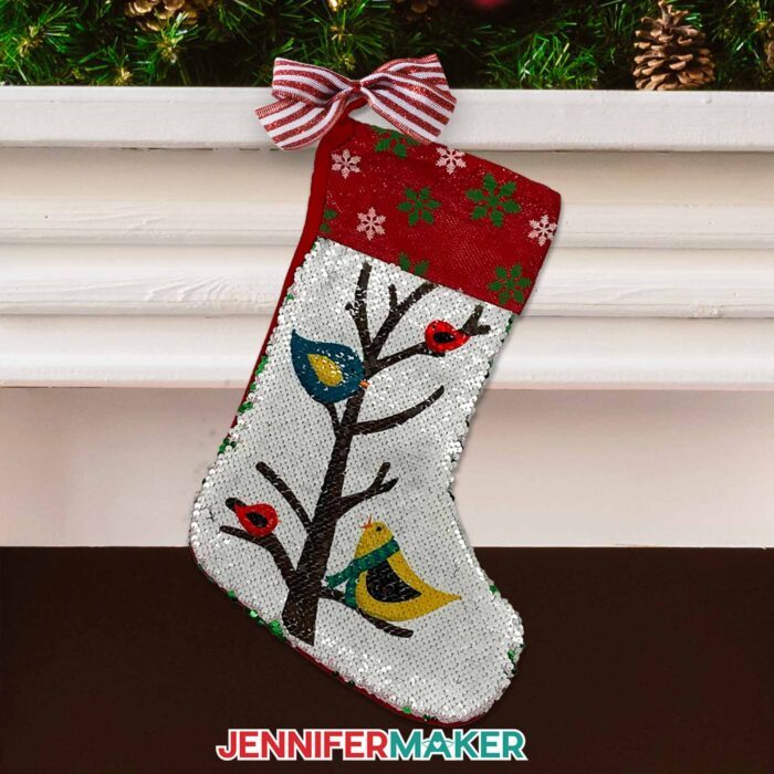 Create a DIY personalized Christmas stocking with sublimation with JenniferMaker's tutorial! A burlap stocking hangs from a white mantle in front of a fireplace. Pinecones and branches decorate the top of the mantle, and the stocking hangs from a striped bow. The stocking is flippable sequins and has multicolored birds on a tree on the front, and green and white snowflakes on the red cuff.