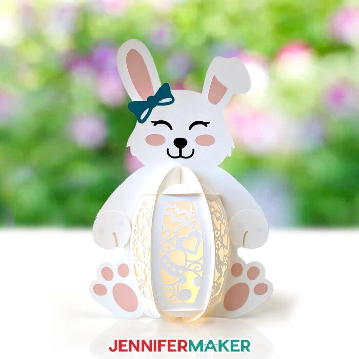 A white cardstock lantern shaped like a bunny sits on a table in a sunny outdoor space with a backdrop of flowers, featuring an intricate cutout design of eggs and hearts through which light emits from an LED tea light. Make your own Easter Lanterns with the free SVG files and tutorial!