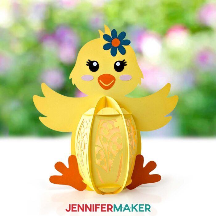 A yellow cardstock lantern shaped like a baby chick sits on a table in a sunny outdoor space with a backdrop of flowers, featuring an intricate cutout design of flowers through which light emits from LED fairy lights. Learn to make Easter Lanterns in the free tutorial!