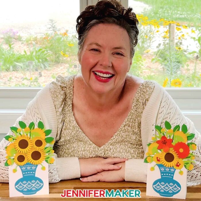 Jennifer Maker smiles, sitting at her craft table and showin off two Easy 3D Bouquet Card projects! One card features sunflowers and one features a mixture of blooms, and both are in an intricate blue paper vase with a built-in stand