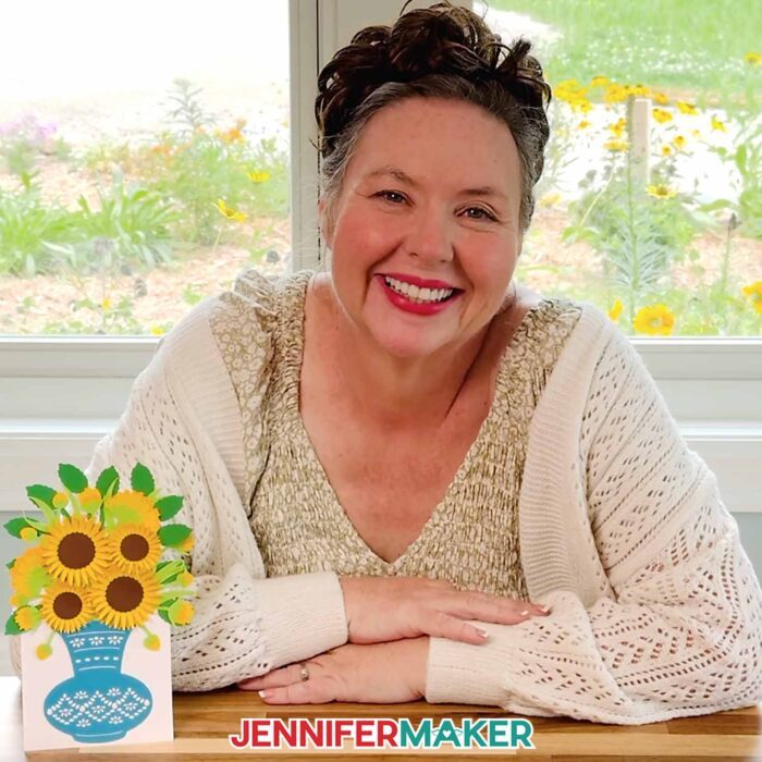 Jennifer Maker smiles, sitting at her craft table, showing off her Easy 3D Bouquet Card! The card features sunflowers in an intricate blue paper vase with a built-in stand