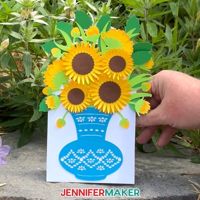 A hand holds the Easy 3D Bouquet Card project against a backdrop of greenery, showing off its pretty paper sunflowers and functional built-in stand