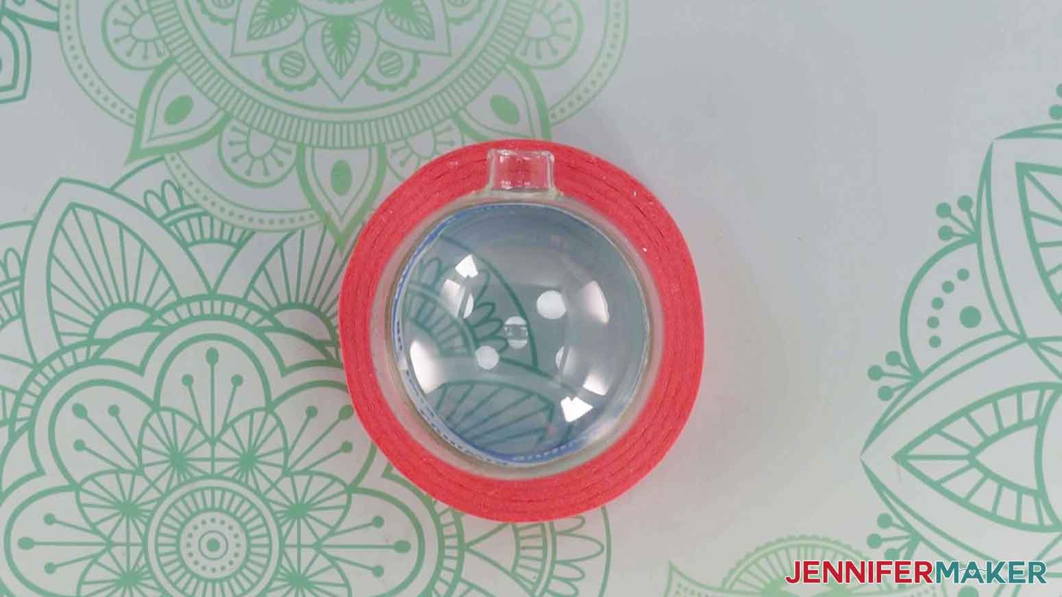 A blank glass ornament placed inside a roll of pink painter's tape lying on its side on a work surface