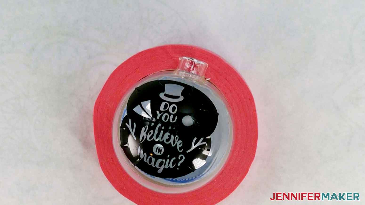 A glass ornament placed in a painter's tape roll with two pieces of black vinyl applied to the surface. The vinyl pieces are overlapping in the center and have a snowman design cut out of them.