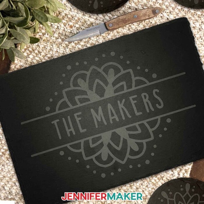 Learn how to etch slate with JenniferMaker's tutorial! Make an etched slate cheeseboard and coasters. Image shows a dark gray slate cheeseboard or charcuterie board, etched with a light gray mandala and "The Makers," sitting on a natural fabric background with etched coasters and a cheese knife nearby.
