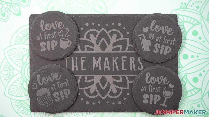 Learn how to etch slate with JenniferMaker's new tutorial! An etched slate cheeseboard with a mandala and "The Makers", flanked by four coasters with cute drink designs and "Love at First Sip" etched onto them. Items are sitting on a mint green cutting mat with mandala designs. 