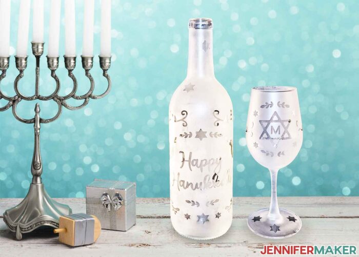 Etched wine glass and a bottle with Hanukkah designs by a Menorah, dreidel, and gift on a table with a blue background.