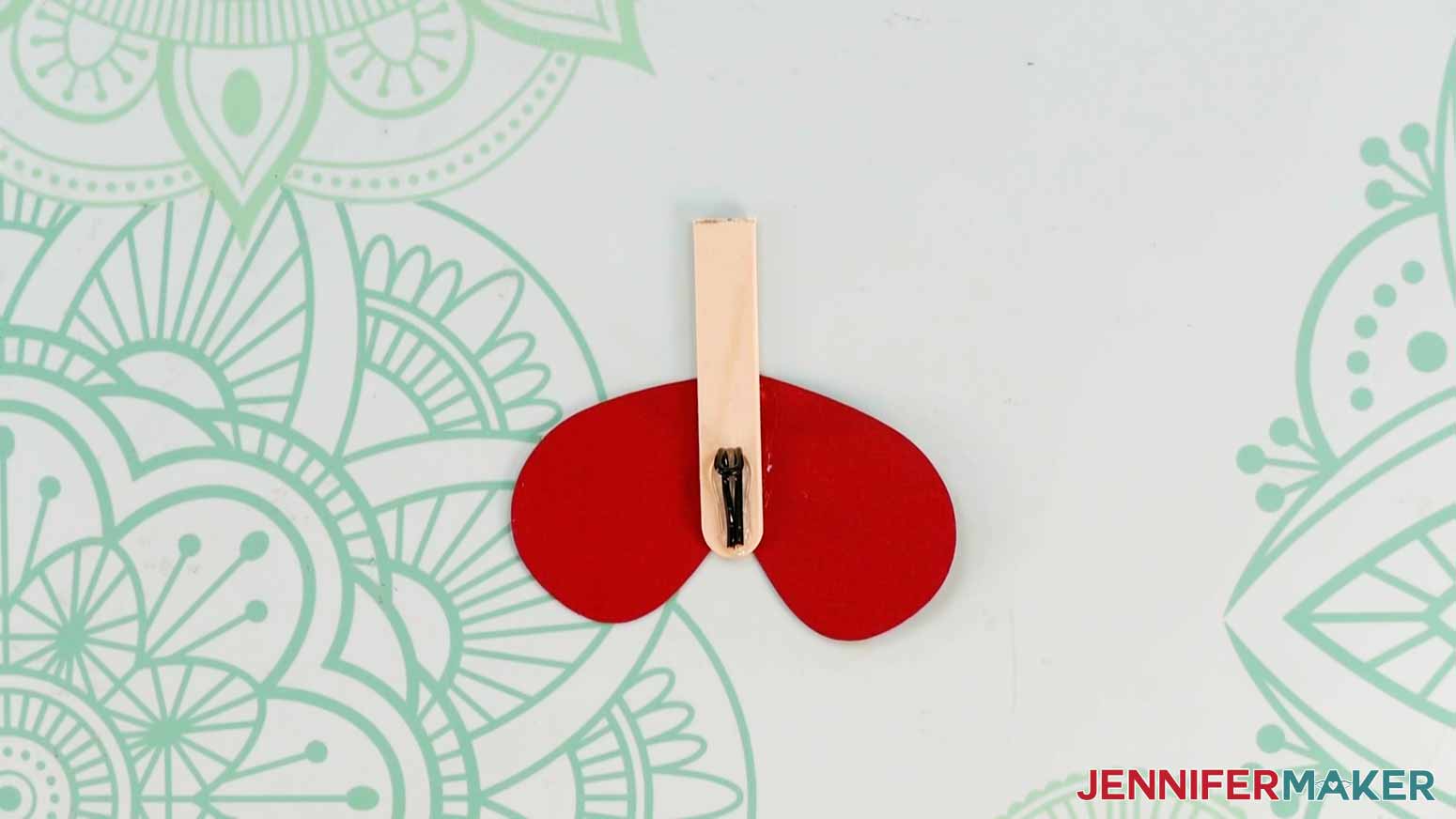 A photo of the bottom part of a magic butterfly for the exploding butterfly box showing a cut popsicle stick with a wire piece glued to the front and a red wing piece attached to the back.