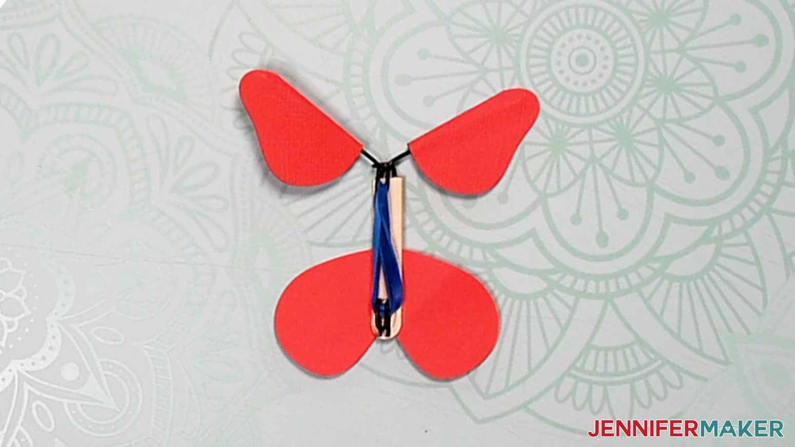 A finished magic butterfly showing red wing pieces on the top and bottom connected with shaped wire pieces attached to a cut popsicle stick. A blue rubber band is looped in the center.