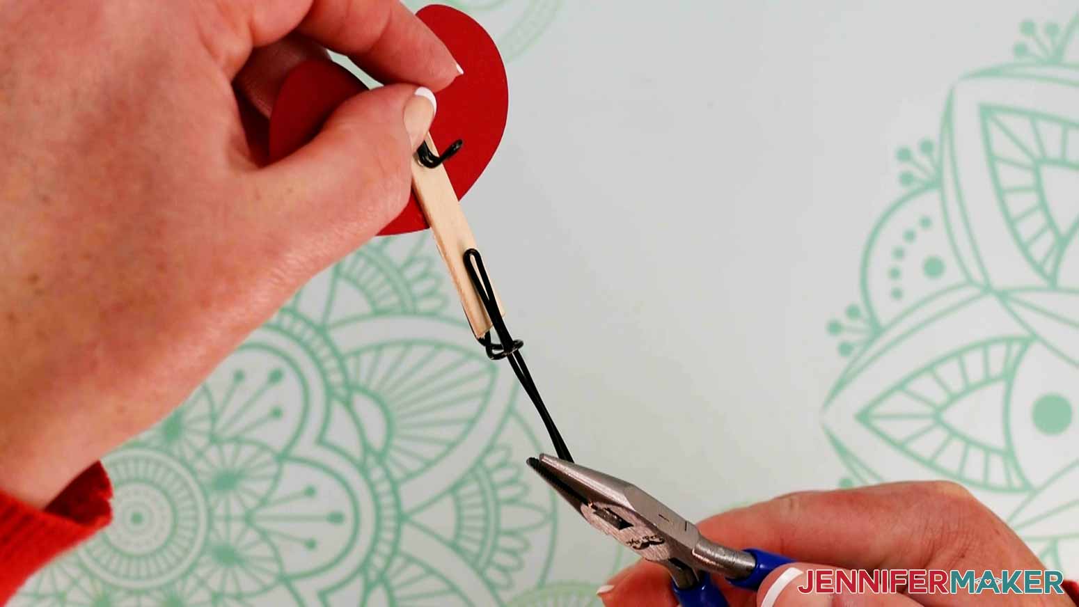 One hand is holding the assembled bottom part of a magic butterfly while another uses needle nosed pliers to hold a shaped piece of wire for the top part. The top wire piece is being shown sliding through the looped wire piece attached to the bottom part of the magic butterfly.