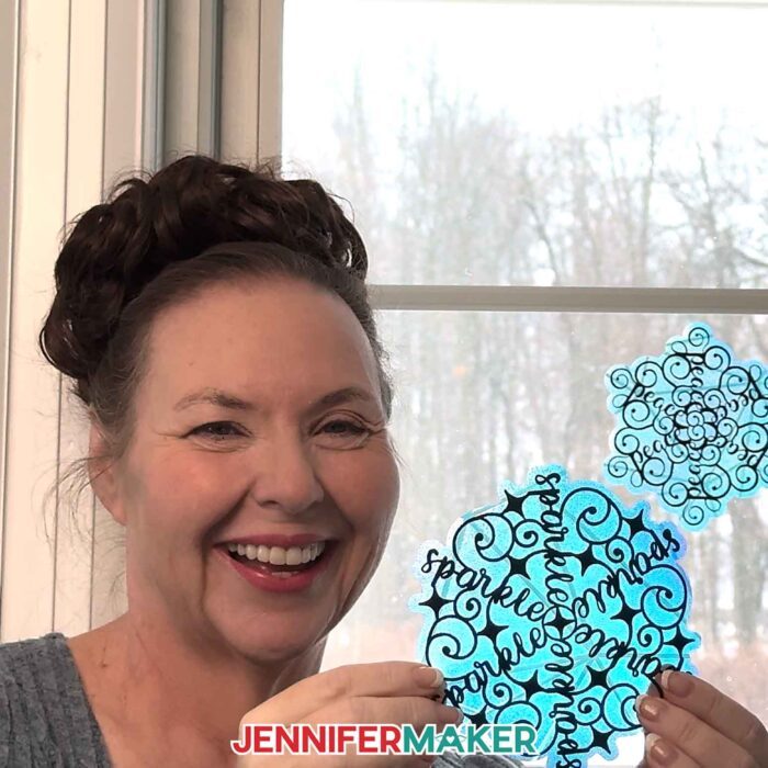 Jennifer Maker, smiling in front of her window looking out onto a snowy Maker Farm, holds up a translucent blue window cling suncatcher decorated with a word made into a Festive Snowflake Design