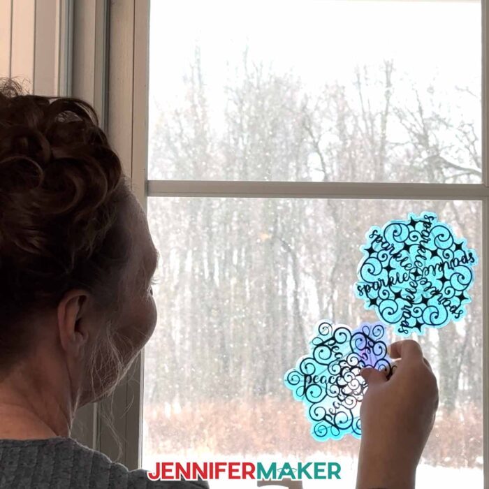 Jennifer Maker, looks out her window onto a snowy Maker Farm, and the window is decorated with two translucent blue window cling suncatchers decorated with a word made into a Festive Snowflake Design