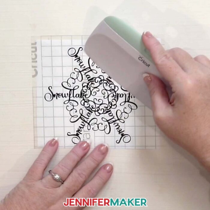 Hands use a scraper to burnish the transfer tape onto the vinyl snowflake design in the Festive Snowflake Design Window Cling tutorial
