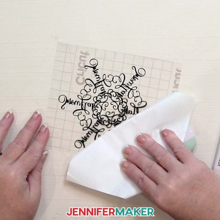 Hands peel the backing off of the transfer tape after applying it to the vinyl snowflake design in the Festive Snowflake Design Window Cling tutorial