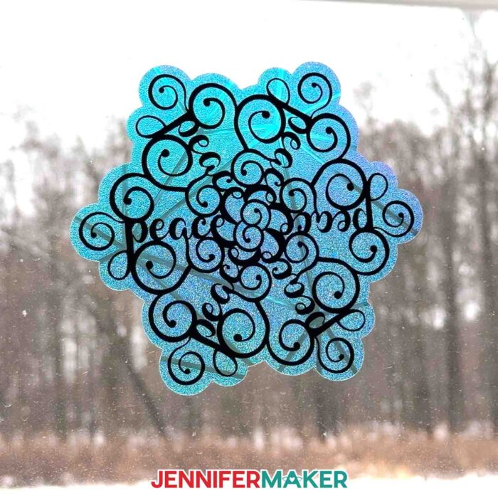 A window from inside the studio at Maker Farm look out onto a snowy landscape -- the window is decorated with a snowflake-shaped window cling with stylized word art saying "Peace" from the Festive Snowflake Designs tutorial
