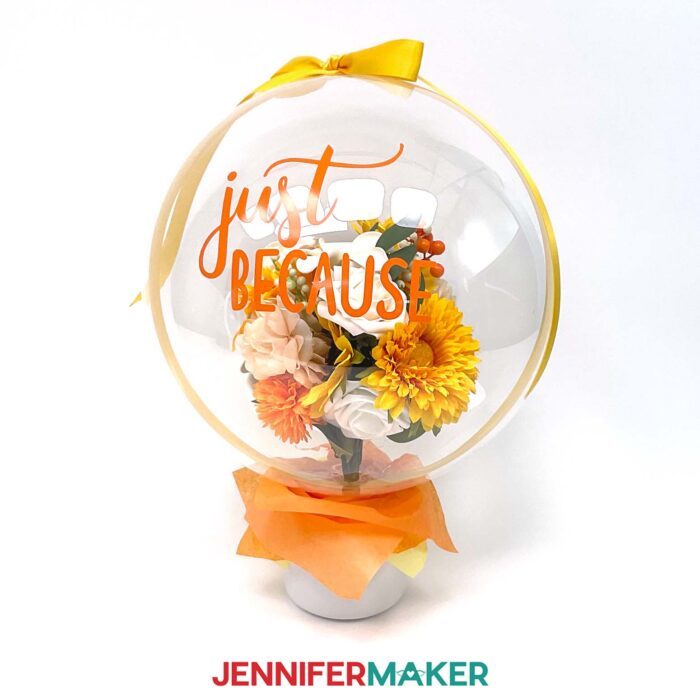 Learn how to put flowers in a balloon with Jennifer Maker's new tutorial! Large flower bouquet on a dark wood tabletop. The balloon reads "Just Because" in yellow vinyl and has yellow and white flowers inside.
