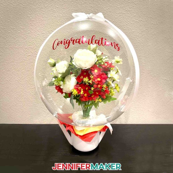Learn how to put flowers in a balloon with Jennifer Maker's new tutorial! Large flower bouquet on a dark wood tabletop. The balloon reads "Congratulations" in red vinyl and has red and white flowers inside.