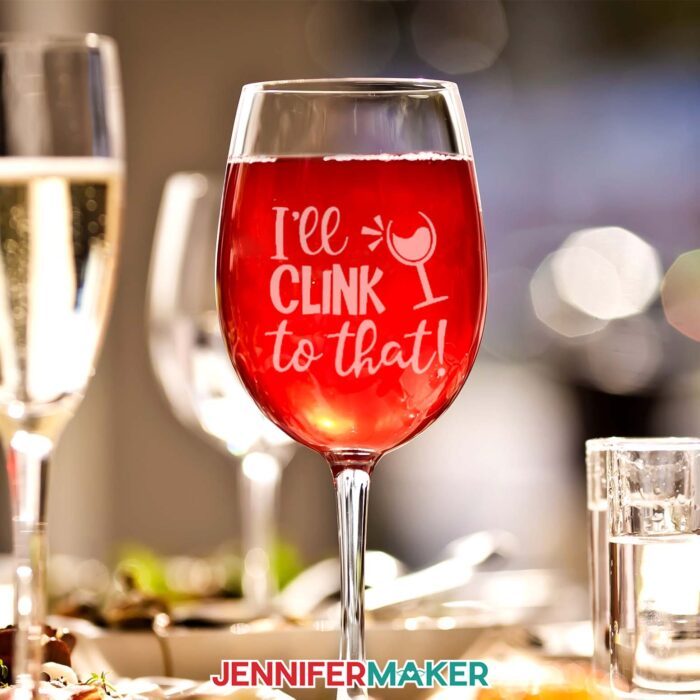 "Etch" glass without etching cream! Learn how with Jennifer Maker's new tutorial. A wine glass sits on a party table, filled with red wine, showing a cute faux etched vinyl decal reading "I'll Clink to That!"