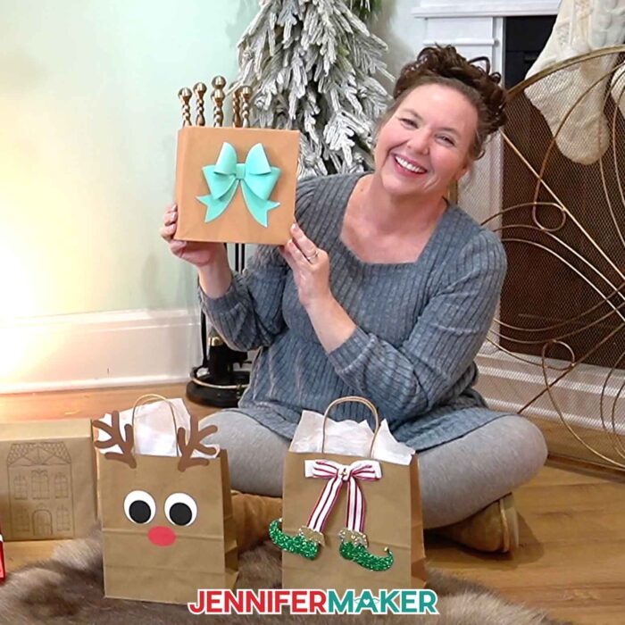 Jennifer sits on the floor behind a reindeer gift bag and an elf gift bag, holding up a box with a big blue cardstock bow on it.