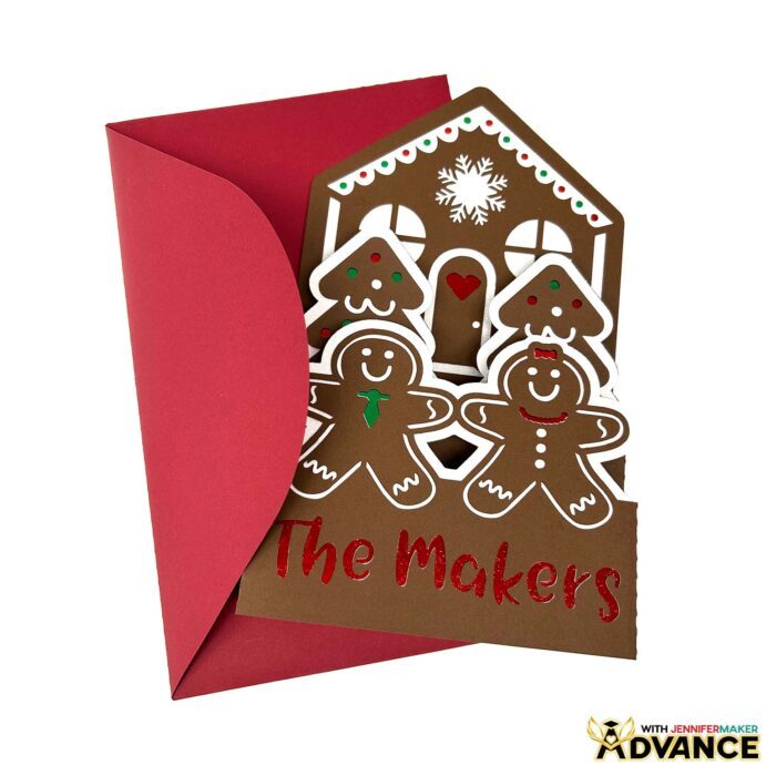 A customized brown Gingerbread House Pop-Up Card sits on display, with red, green, and white details and the name "Maker" cut out of the front, with a matching red envelope