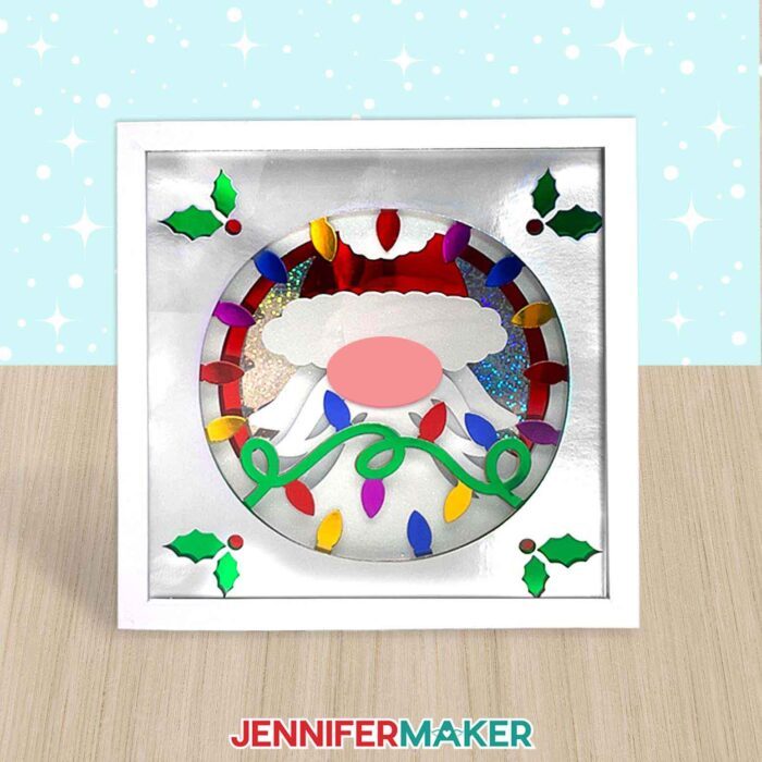 A gnome shadow box with Christmas lights and holly made from layered foil cardstock sits on a table.