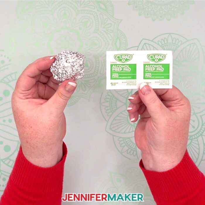 Jennifer Maker shows a ball of aluminum foil in one hand and isopropyl alcohol wipes in the other, both used to clean the Fine Point Blade in a Cricut cutting machine, demonstrated in the Gnome Gift Set tutorial
