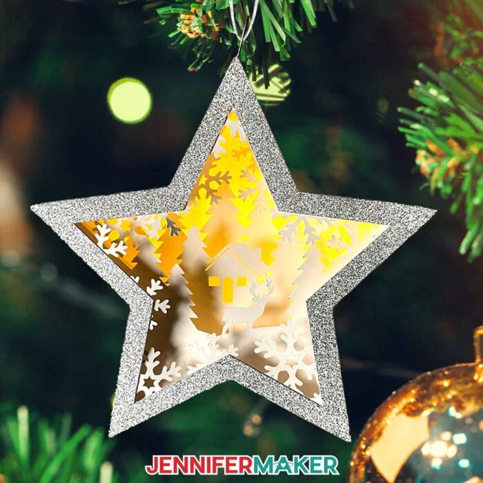 Make DIY Hanging Star Lanterns with JenniferMaker's tutorial! A miniature star-shaped layered cardstock ornament with a winter cottage scene with snowflakes and a deer inside hangs on a Christmas tree with a backdrop of twinkling lights.