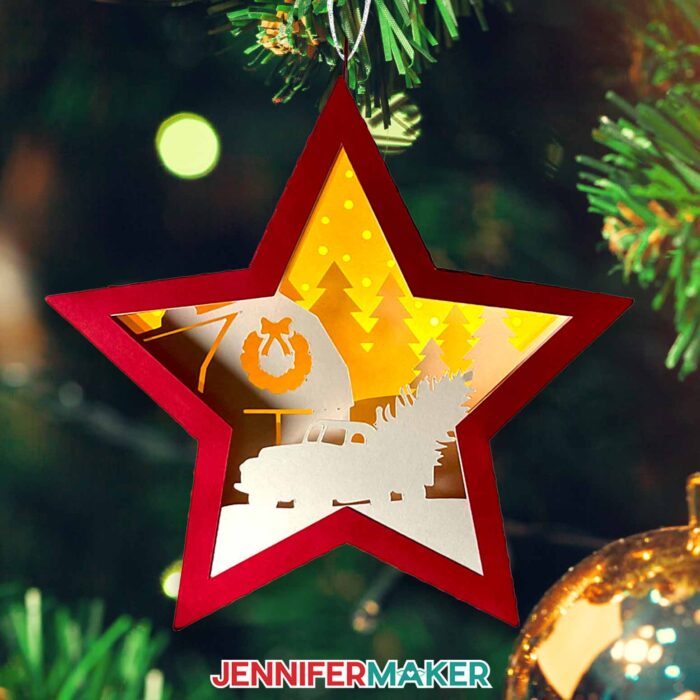 Make DIY Hanging Star Lanterns with JenniferMaker's tutorial! A miniature star-shaped layered cardstock ornament with a winter truck scene with pine trees and a barn with a wreath inside hangs on a Christmas tree with a backdrop of twinkling lights.