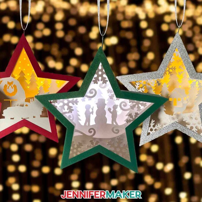 Make DIY Hanging Star Lanterns with JenniferMaker's tutorial! Three miniature star-shaped layered cardstock ornaments with winter and holiday scenes inside, hanging on a backdrop of twinkling Christmas lights. Make Cricut Christmas Ornaments with JenniferMaker's tutorial!