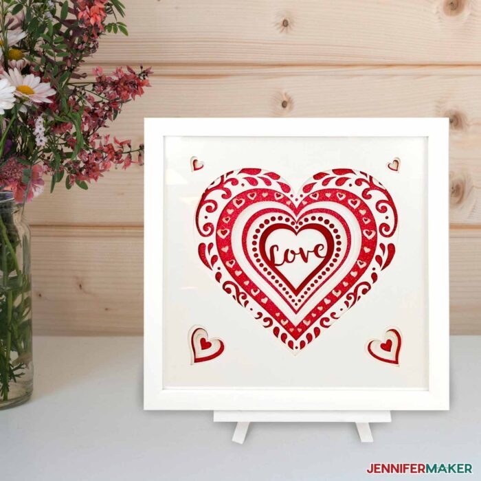 "Love" in a layered heart of white and red cardstock in a white frame on a table with flowers