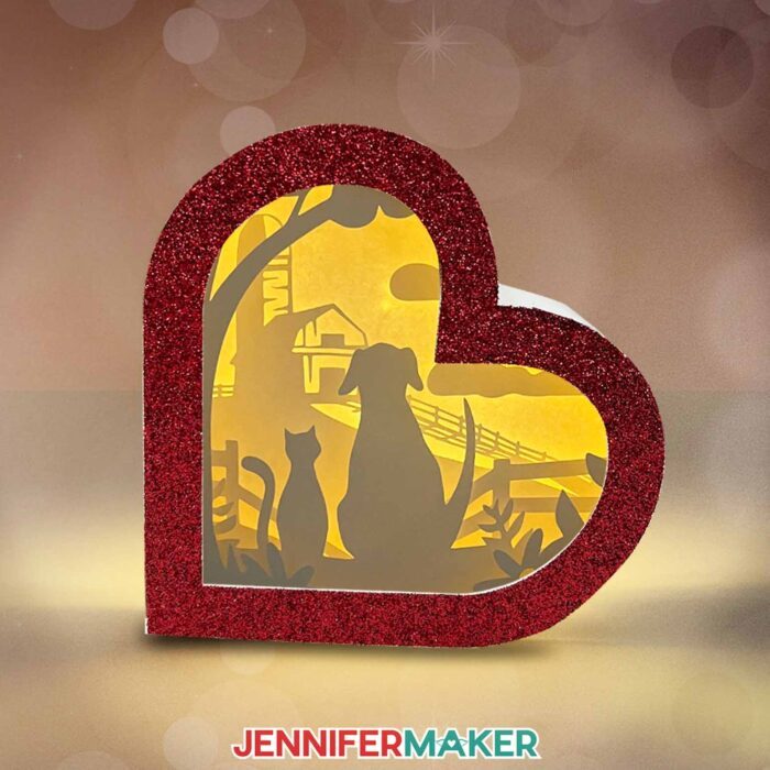 Make a Heart-Shaped Shadow Box with Jennifer Maker's tutorial! A heart-shaped shadow box with red glitter frame and a scene with a dog and cat overlooking a barn.