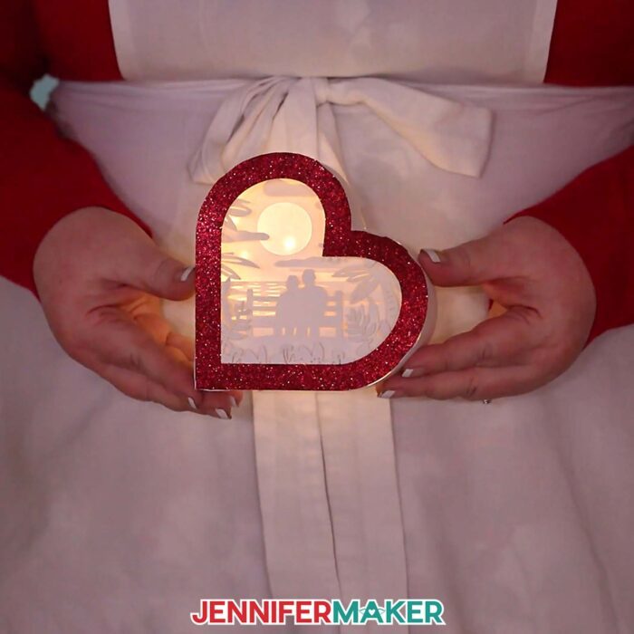 Make a Heart-Shaped Shadow Box with Jennifer Maker's tutorial! Jennifer is holding an illuminated heart-shaped shadow box with red glitter frame and a scene with a couple on a bench overlooking an ocean sunset.