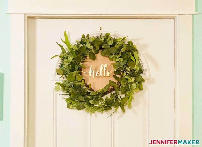 Faux Eucalyptus wreath with a gold hello cutout on posterboard in the center and hanging on a white door