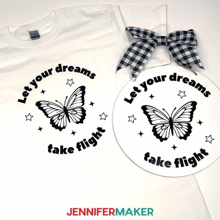 A white T-shirt and a light colored wood sign lie on a surface decorated with black vinyl butterfly decals that say "Let your dreams take flight." Follow the Learn How To Create A Cut File In Canva tutorial to learn how to make your own custom vinyl designs