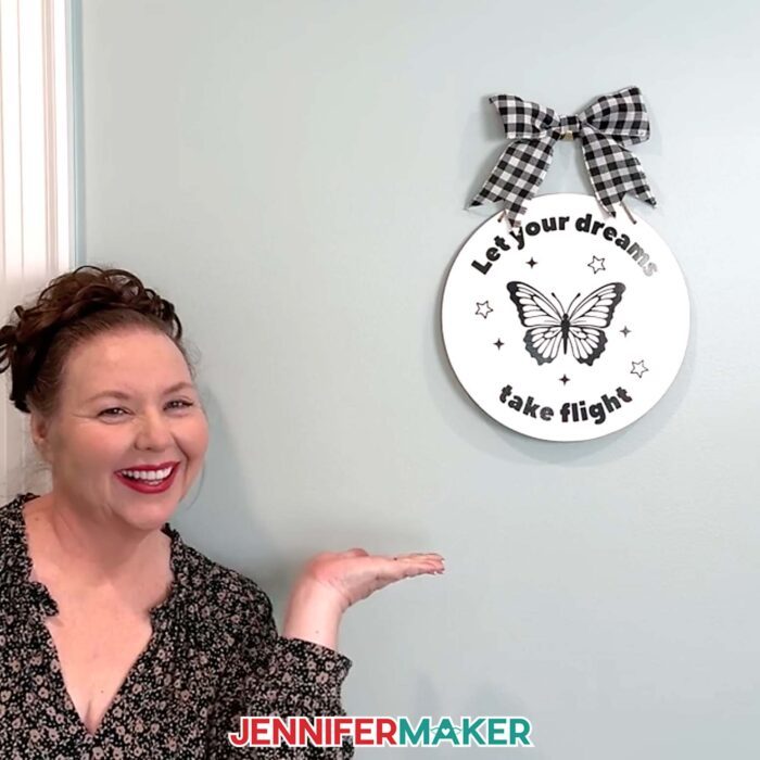 Jennifer Maker shows off a round sign decorated with a butterfly decal that says "Let your dreams take flight" that she made in the Learn How To Create A Cut File In Canva tutorial