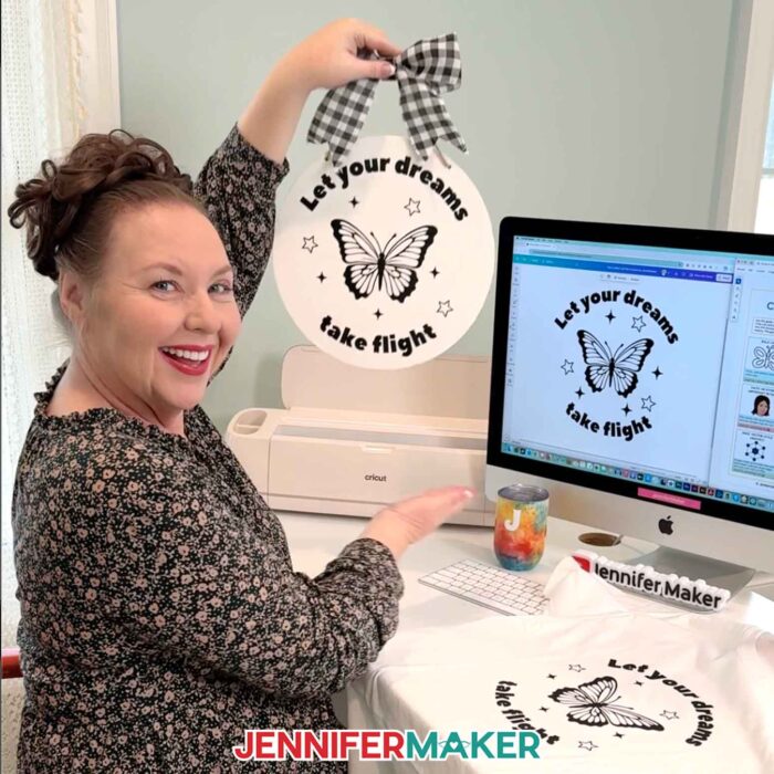 Jennifer Maker sits at her computer and shows off a round sign decorated with a butterfly decal that says "Let your dreams take flight" that she made in the Learn How To Create A Cut File In Canva tutorial