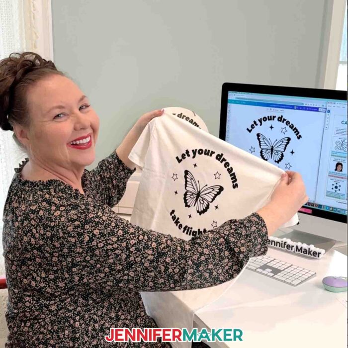 Jennifer Maker sits at her computer and shows off a T-shirt decorated with a butterfly decal that says "Let your dreams take flight" that she made in the Learn How To Create A Cut File In Canva tutorial