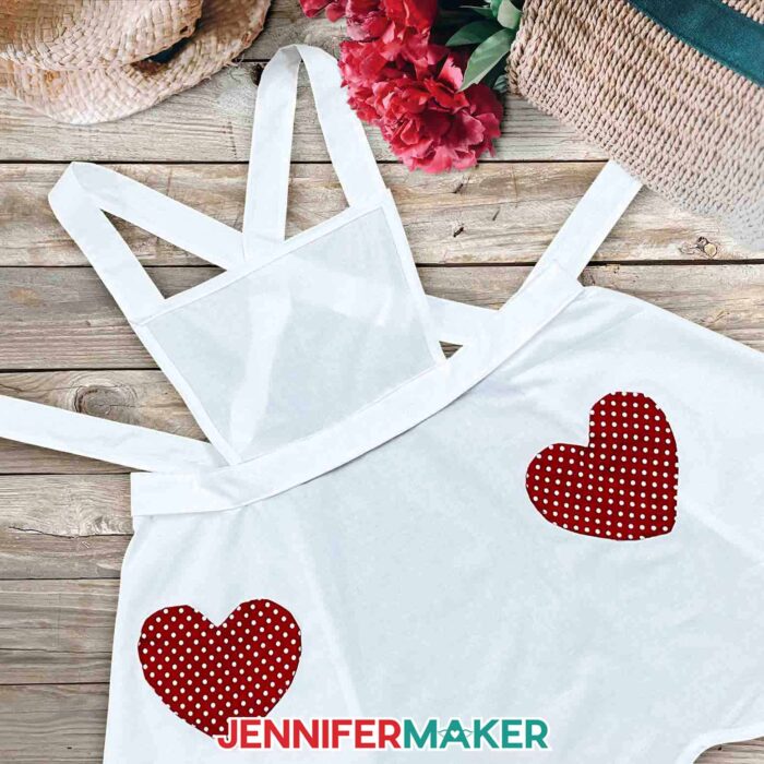 A white vintage style apron with red heart-shaped pockets and cross back straps laid on a wood background.