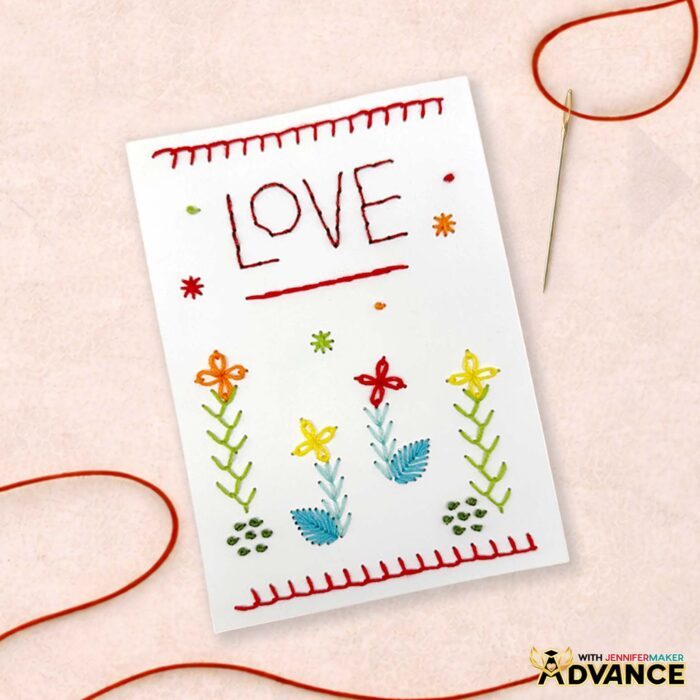 A 5 by 7 card with a custom design using stitches from the stitch book: The word 'love' is backstitched across the top half in red, with orange, red, and yellow daisy flowers stitched across the bottom half.