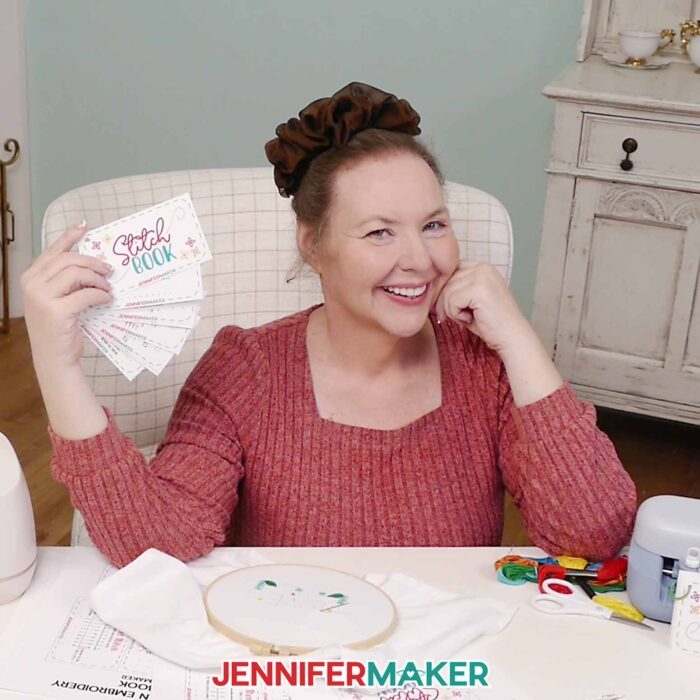 Jennifer sits at her craft desk, surrounded by sewing supplies, holding up her stitch book with the cards fanned out.