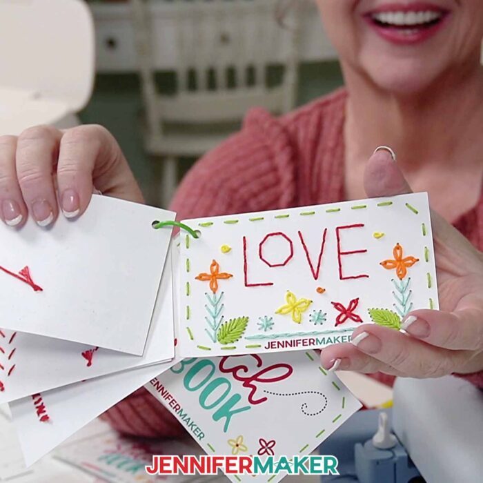 Jennifer holds up her stitched practice design card. It has 'love' back stitched in red, with colorful embroidered flowers and accent stitches surrounding it.