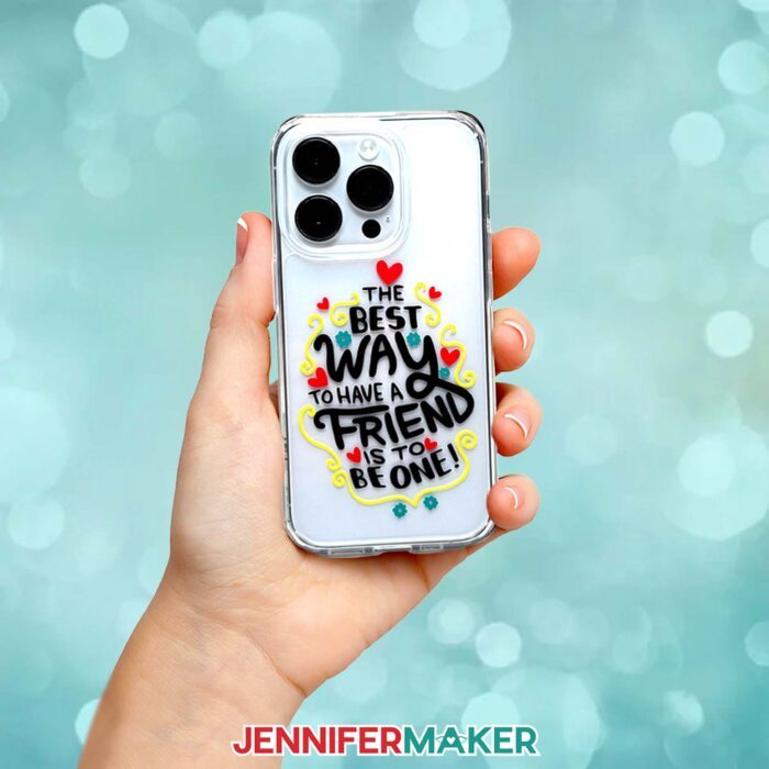 Learn how to make a phone case with vinyl decals in JenniferMaker's tutorial! A hand holds a smartphone with a plastic case. The case has "The best way to have a friend is to be one!" in black vinyl surrounded by vinyl red hearts, teal flowers, and yellow swirls.