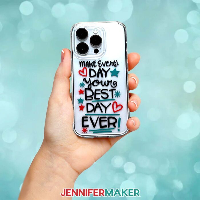 Learn how to make a phone case with vinyl decals in JenniferMaker's tutorial! A hand holds a smartphone with a plastic case. The case has "Make every day your best day ever!" in black vinyl surrounded by hearts and stars in red and teal vinyl.