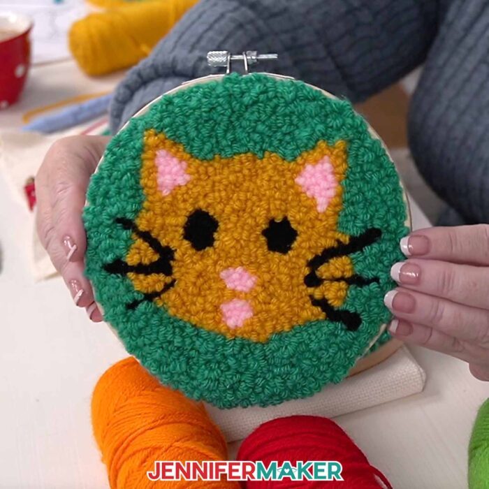 A close-up view of a cat face punch needle project for beginners. The orange cat has black whiskers and eyes; pink ears, nose, and mouth; and a teal background.