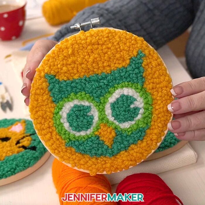 A close-up look of the owl face punch needle project: the owl has a teal face with green eyes and an orange beak on an orange background.