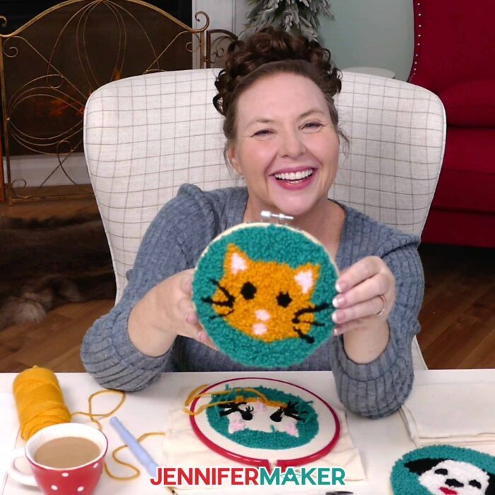 Jennifer sits at her desk holding up her cat face punch needle project.
