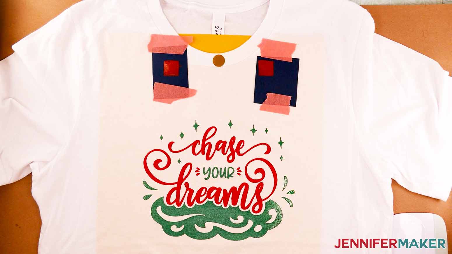 A white screen printed shirt with the Chase Your Dreams design in red and emerald ink. Two small pieces of cardstock with red inked squares are taped to the top of the shirt. A vinyl circle is also placed on the center of the collar.