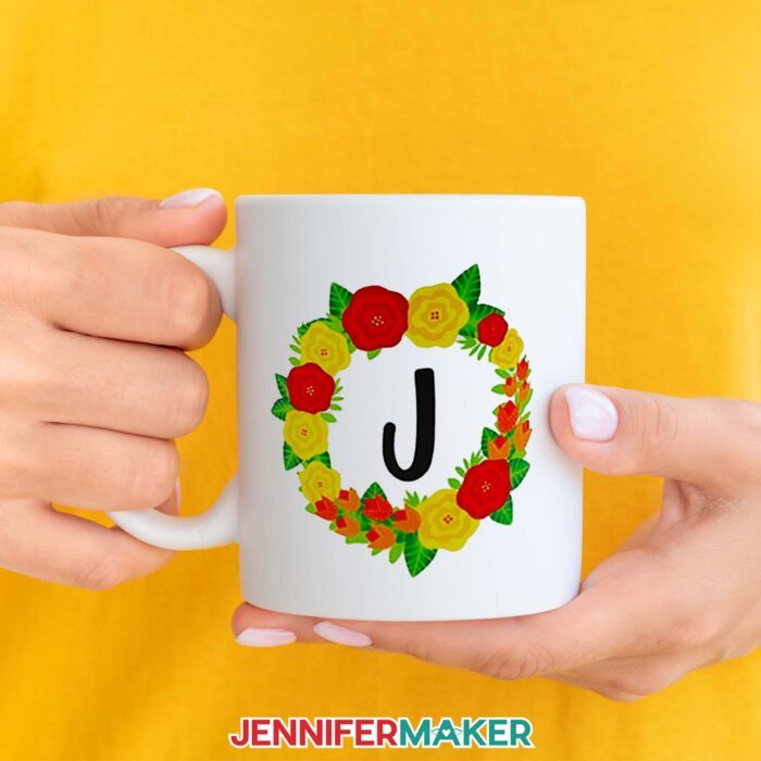 Sublimated floral mug, held against a background of yellow. A wreath design encircles a bold "J" and is made up of red, orange, and yellow flowers. Learn how to sublimate mugs with JenniferMaker's tutorial!